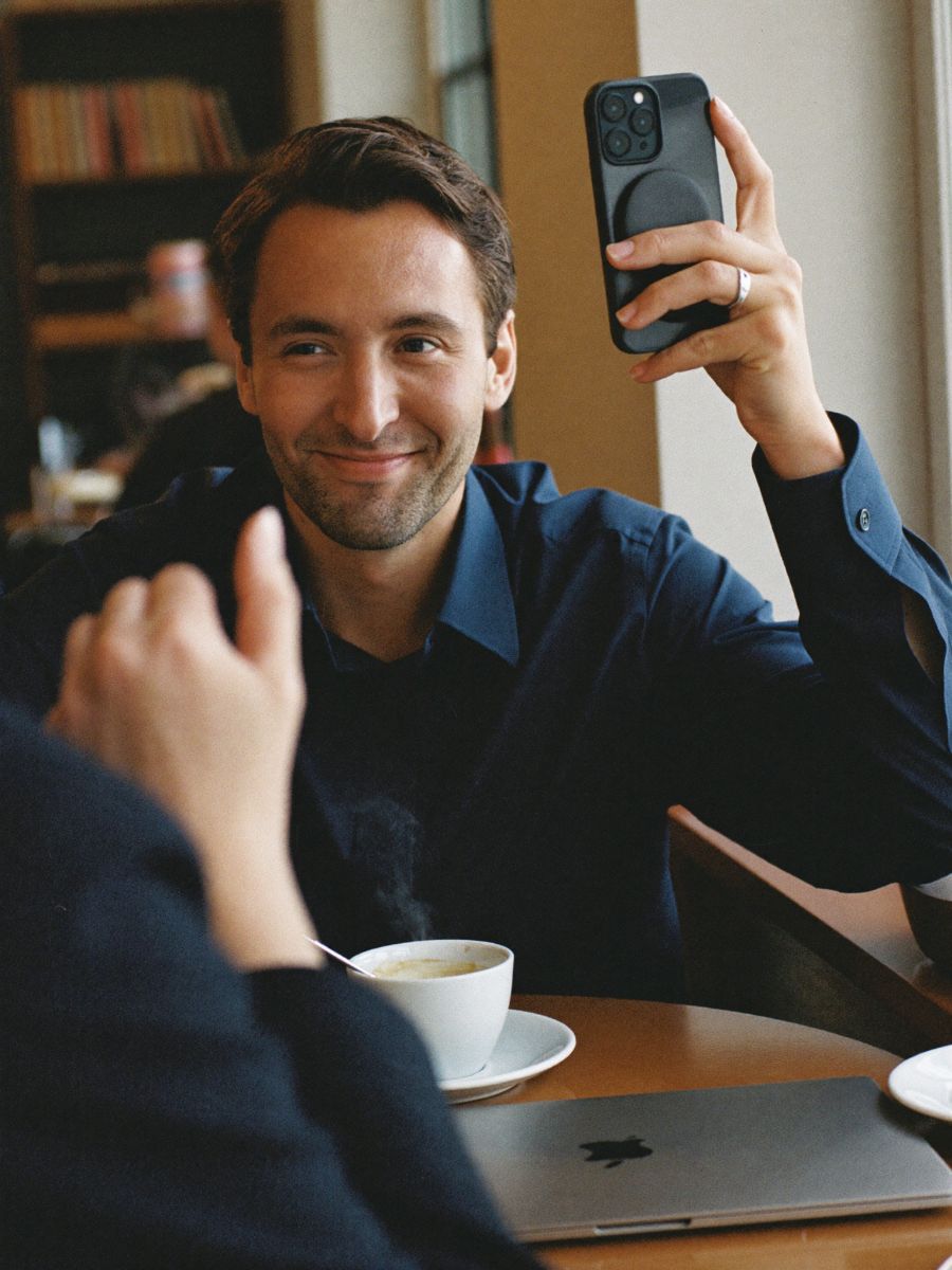 A smiling sales manager in a navy shirt at a café table, holding up his phone to a client
