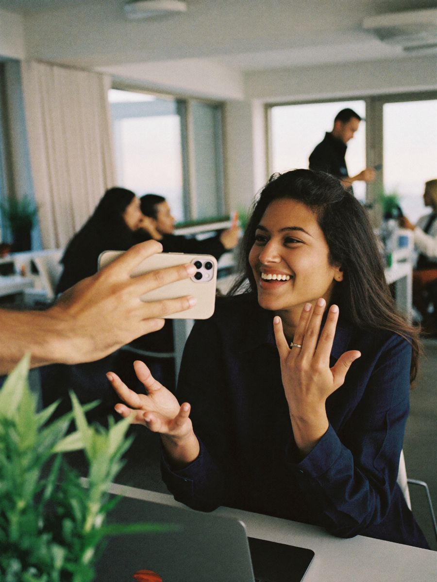A new colleague holding out his phone to a smiling coworker at her desk in an open-plan office