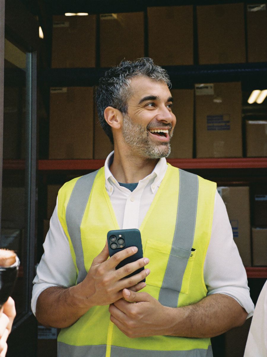 An operations manager in a hi-vis vest in a warehouse office, holding his phone and smiling